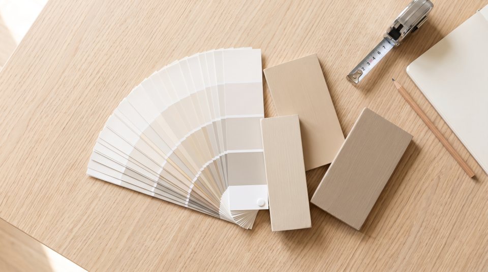 An overhead view of a fanned-out paint color deck, painted sample boards, a tape measure and a notebook on a pale oak table
