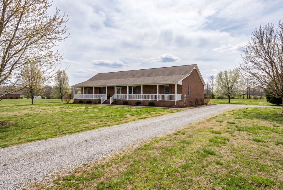 A brick ranch house with a long white porch at the end of a gravel drive